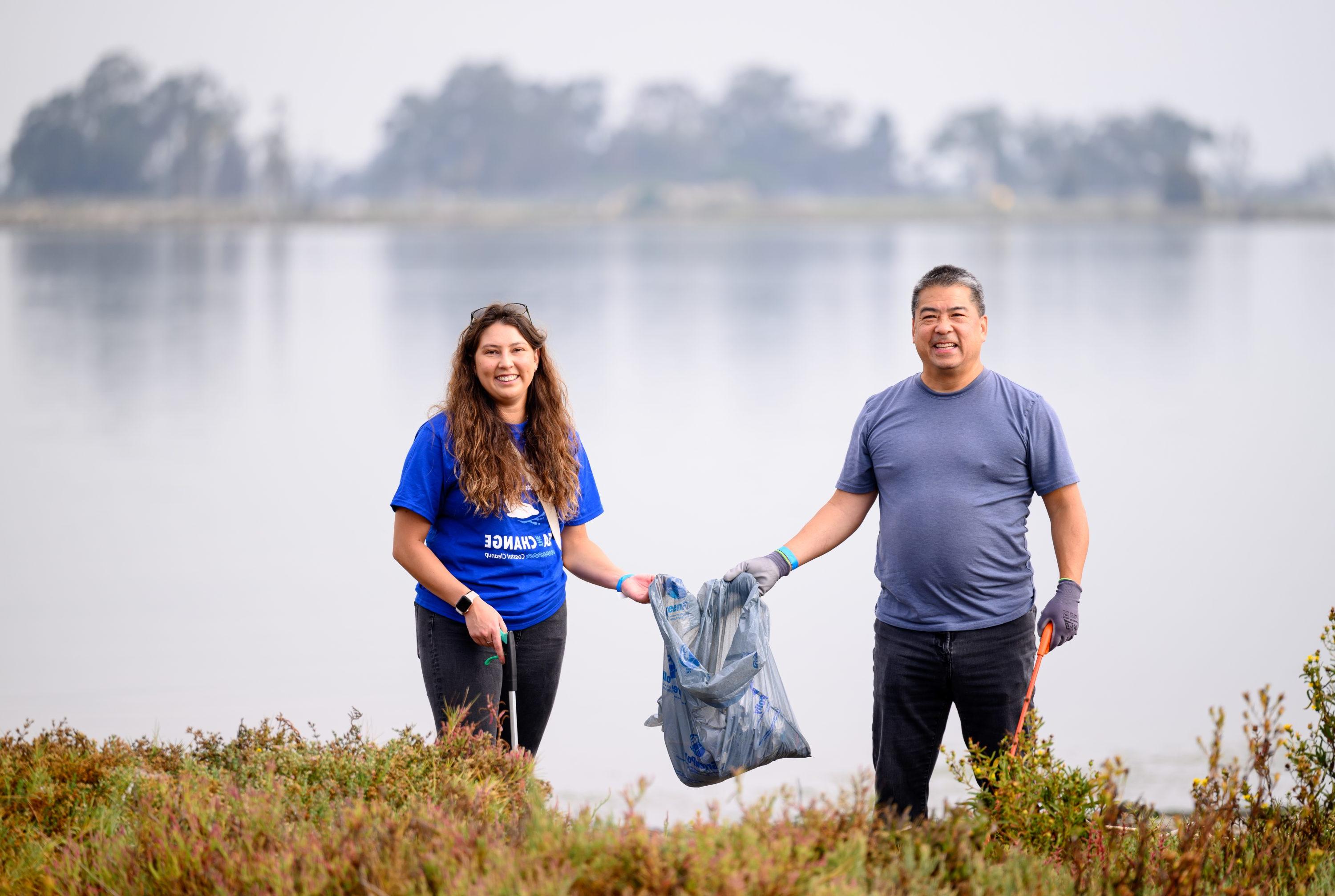 Two volunteers hold one trash bag and hold their grabbers with the Oakland Estuary in the background. 